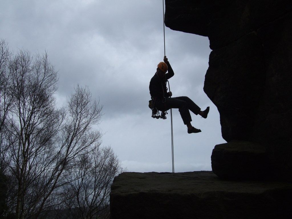 A picture of Jesse abseiling off the top of a gritstone pinnacle in the Peak District. He is in silhouette against a dark grey sky.