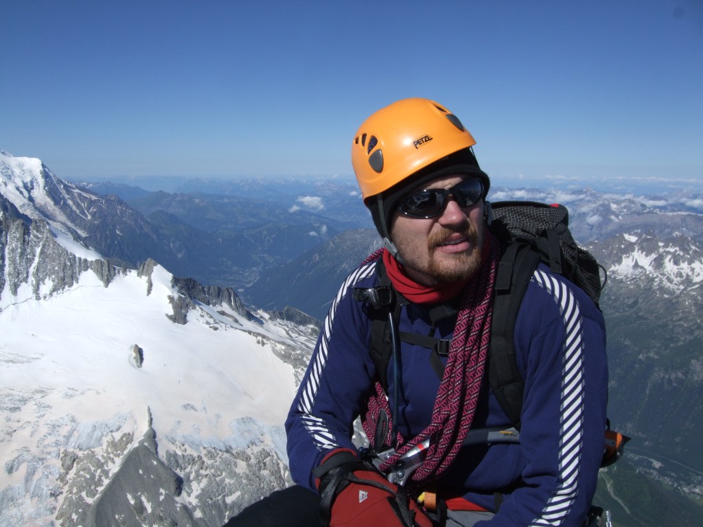 A picture of Jesse sat at the top of a mountain in the alps with snowy peaks in the background. He is wearing an orange helmet, has a rope coiled over his shoulder and is holding an ice axe.