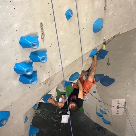 A picture of Jesse competing in an indoor climbing competition. He is climbing a very overhanging roof, making a long reach out to a blue volcano style hold, with his heel resting on a volume.