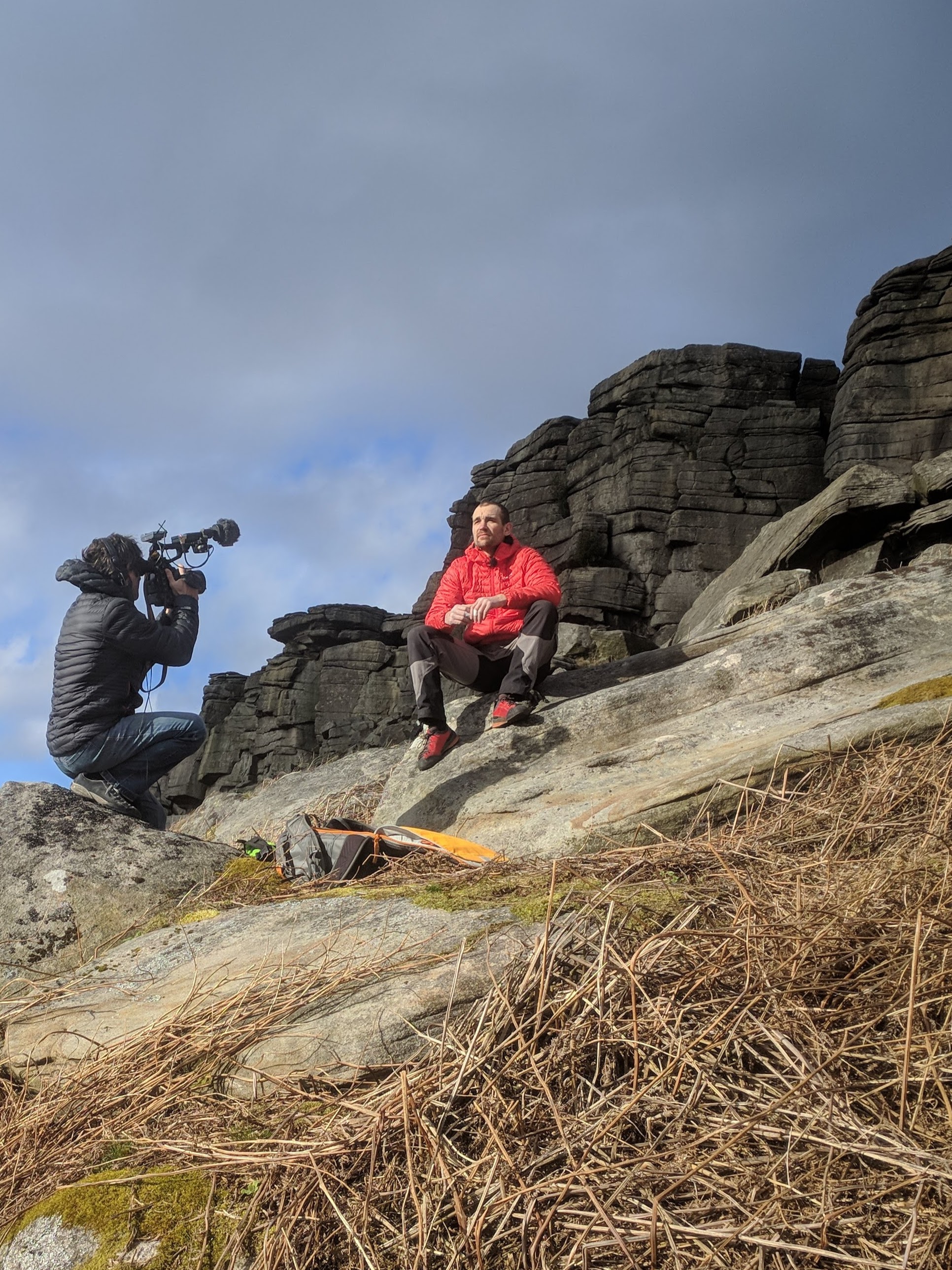 A picture of Jesse sat on a rock wearing his red jacket in the centre of the picture. Alastair is squatting to one side of him with his big camera filming an interview. The gritstone crag of Stanage popular is in the background.