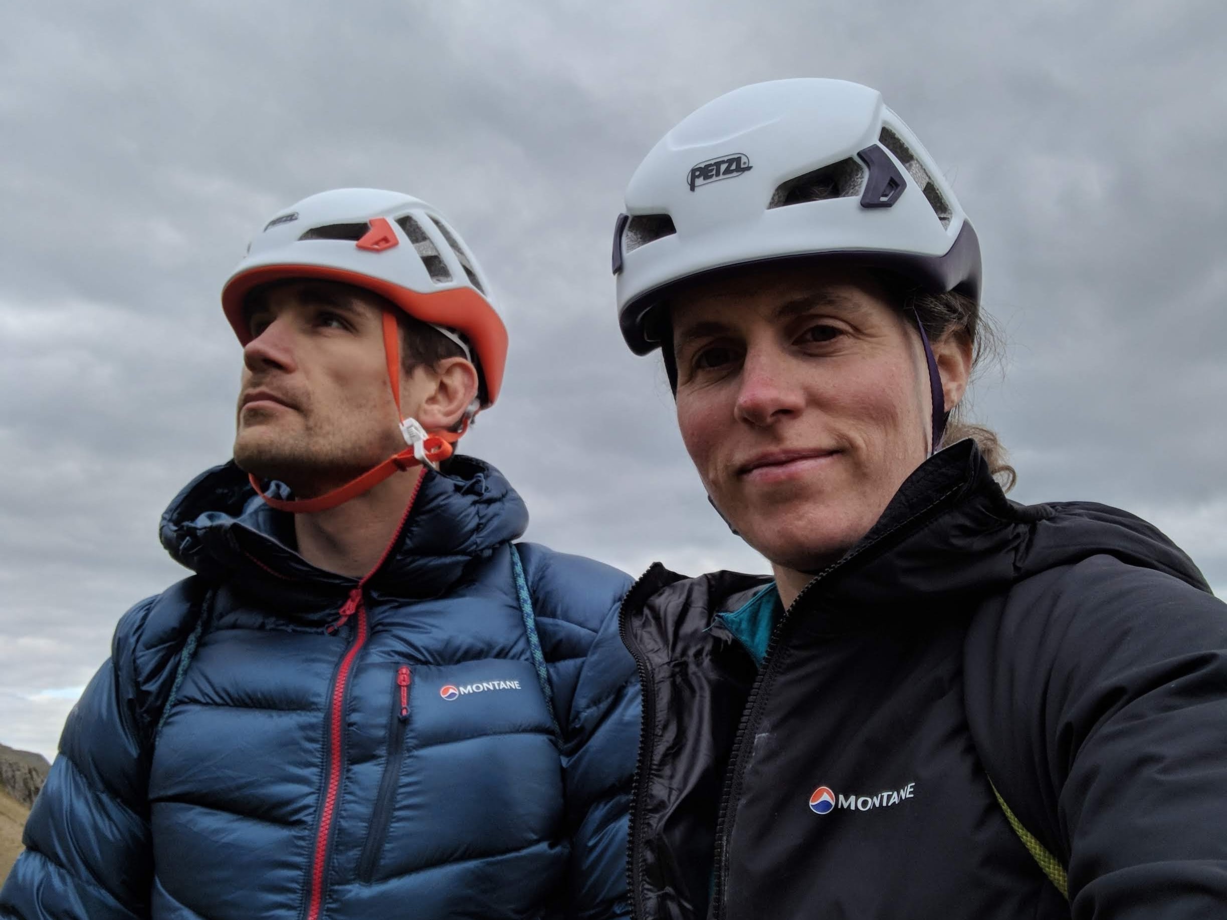 A picture of Jesse and Molly, both wearing Petzl climbing helmets and Montane jackets. Molly is looking at the camera and Jesse is looking out into the distance. They are wearing coiled ropes on their backs as the approach the crag.