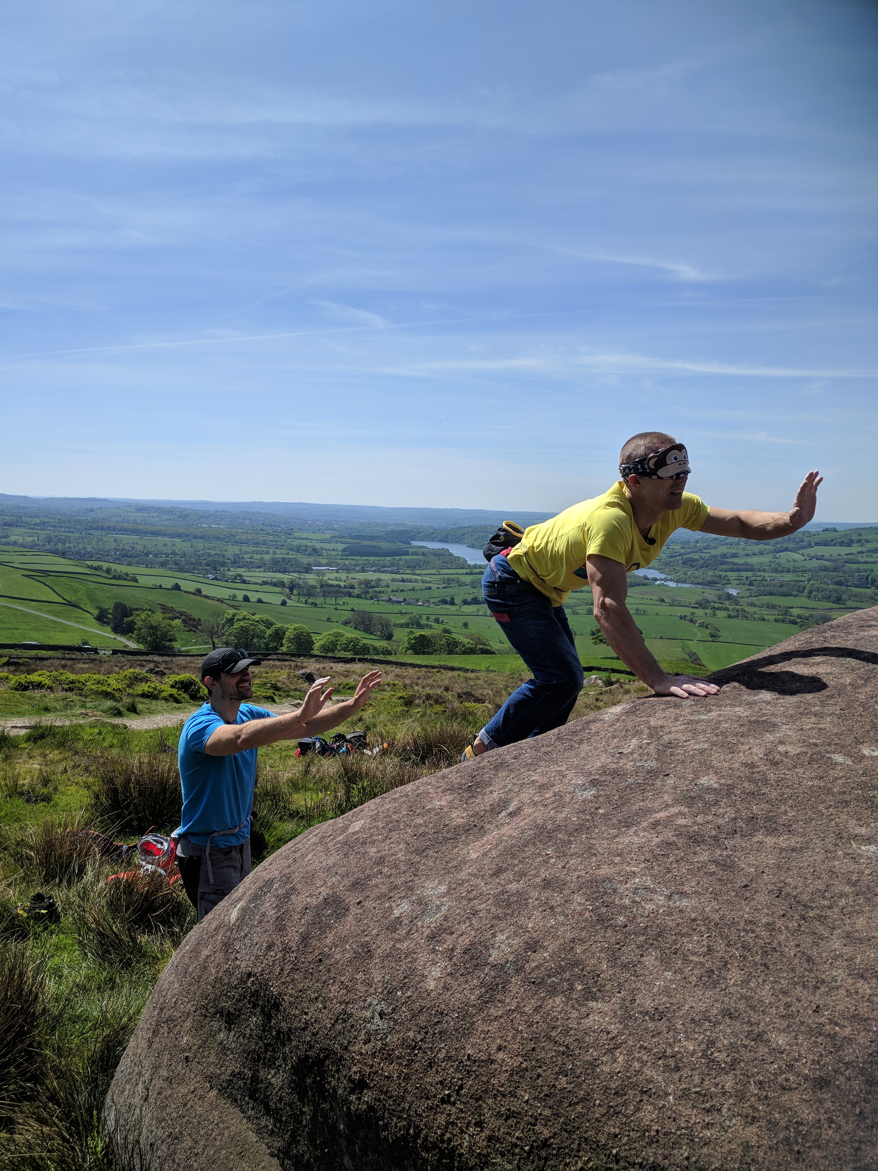 A picture of Jesse and Neil bouldering at the roaches. Jesse is stood at the base of the boulder with both arms in the air, spotting neil. Neil is wearing a monkey eye mask and is reaching out into midair trying to search for the next hold. He is squatting down and reaching forward in an extremely funny pose.
