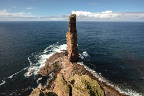 A picture of the Old Man of Hoy taken from the headland looking out to sea. The sea stack is 137 meters tall and is standing proud right in the middle of the picture. Waves are crashing around its base and it is a beautiful blue sky day.