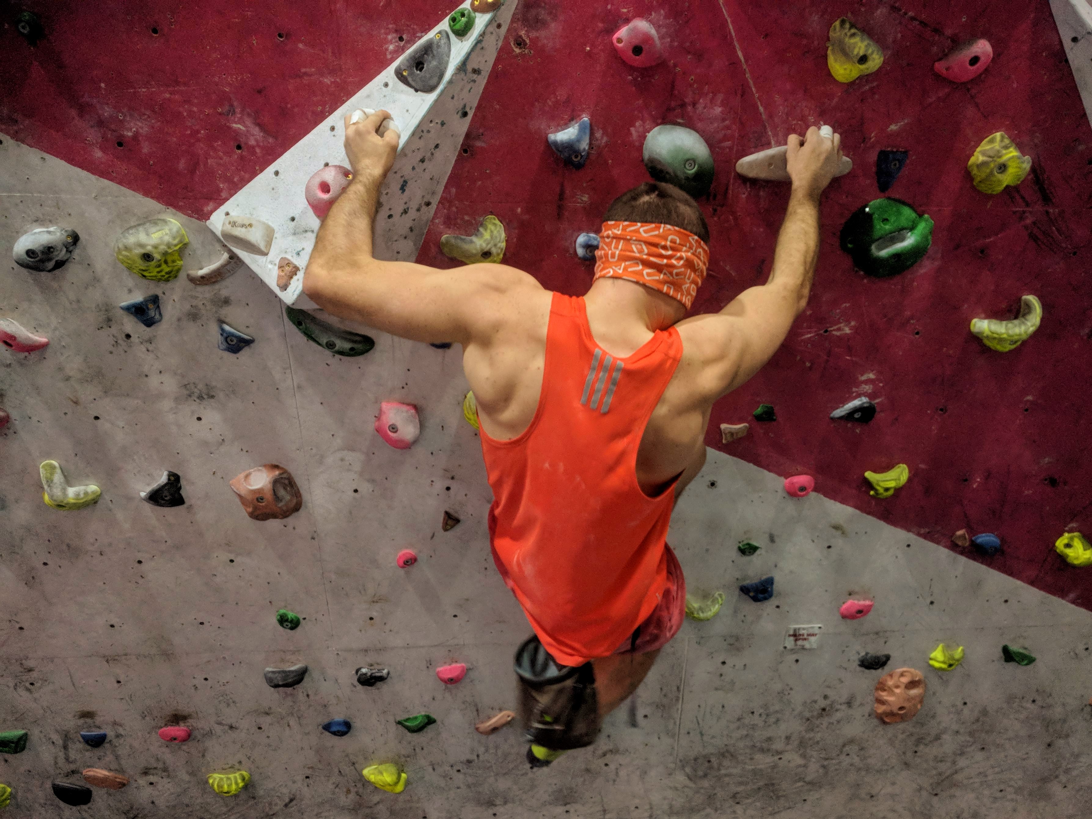A picture of Jesse wearing an orange vest and an orange blindfold. He is climbing on the wooden holds on the steep circuit board at his local climbing wall.