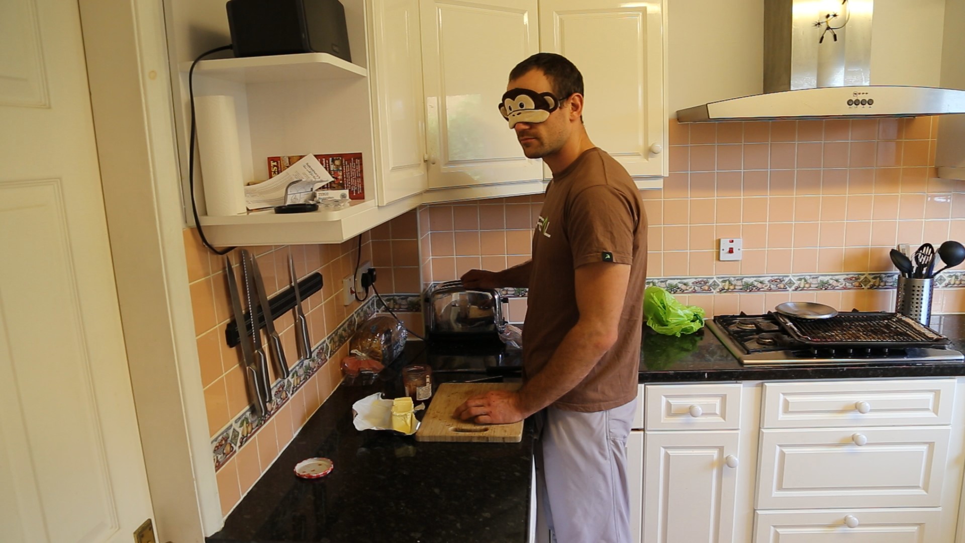 A picture of Jesse in his kitchen, he is reaching for the toaster with butter and jam at the ready. He is wearing a monkey eye mask.