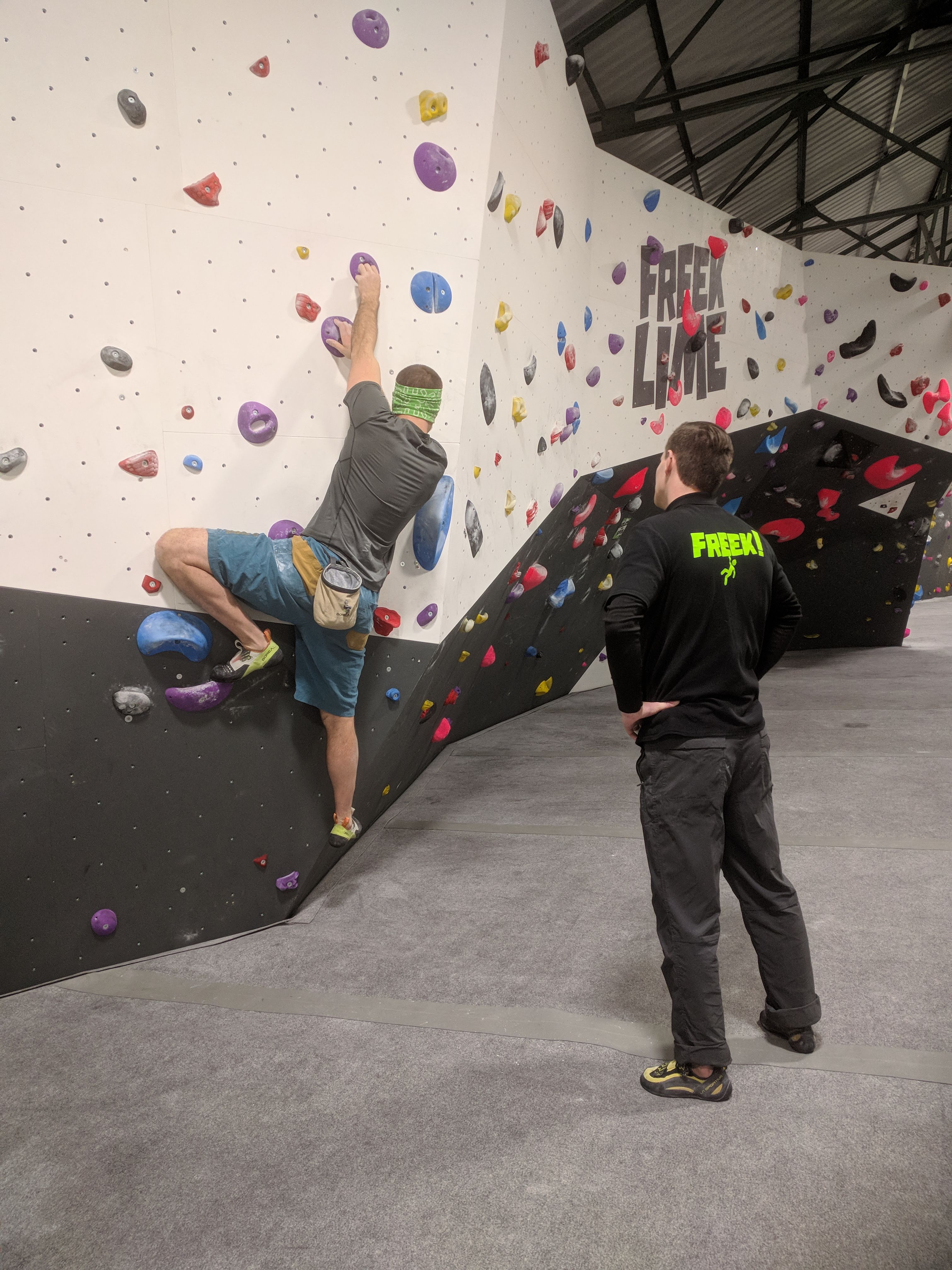 A picture of Jesse climbing a boulder problem with purple pockety holds. A member of staff is stood on the mat behind Jesse acting as his sight-guide.
