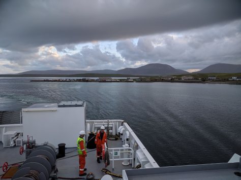 A picture taken from onboard the ferry to Hoy. The back of the ferry is visible with sea, islands and mountains in the background.