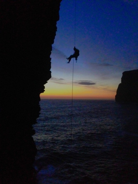 A picture of Jesse on the last free hanging abseil to get down to the base of the sea stack. The sun is setting and he and the stack are silhouetted in the foreground. It is very nearly dark.