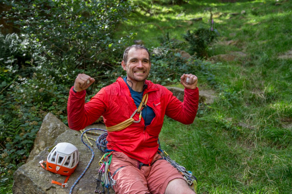 A picture of Jesse sat on a rock at the base of the crag wearing a bright red jacket. He has both fists clenched and his arms in the air and a huge grim on his face. He looks extremely pleased to have successfully climbed the route.