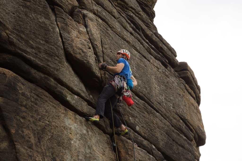 A picture of Jesse rock climbing. He is wearing a blue t-shirt, a white helmet and has a harness full of gear dangling around his waist. He is hanging on to a small side pull hold with his left hand, with two feet in a horizontal break. His right hand is dipping in his chalk bag.