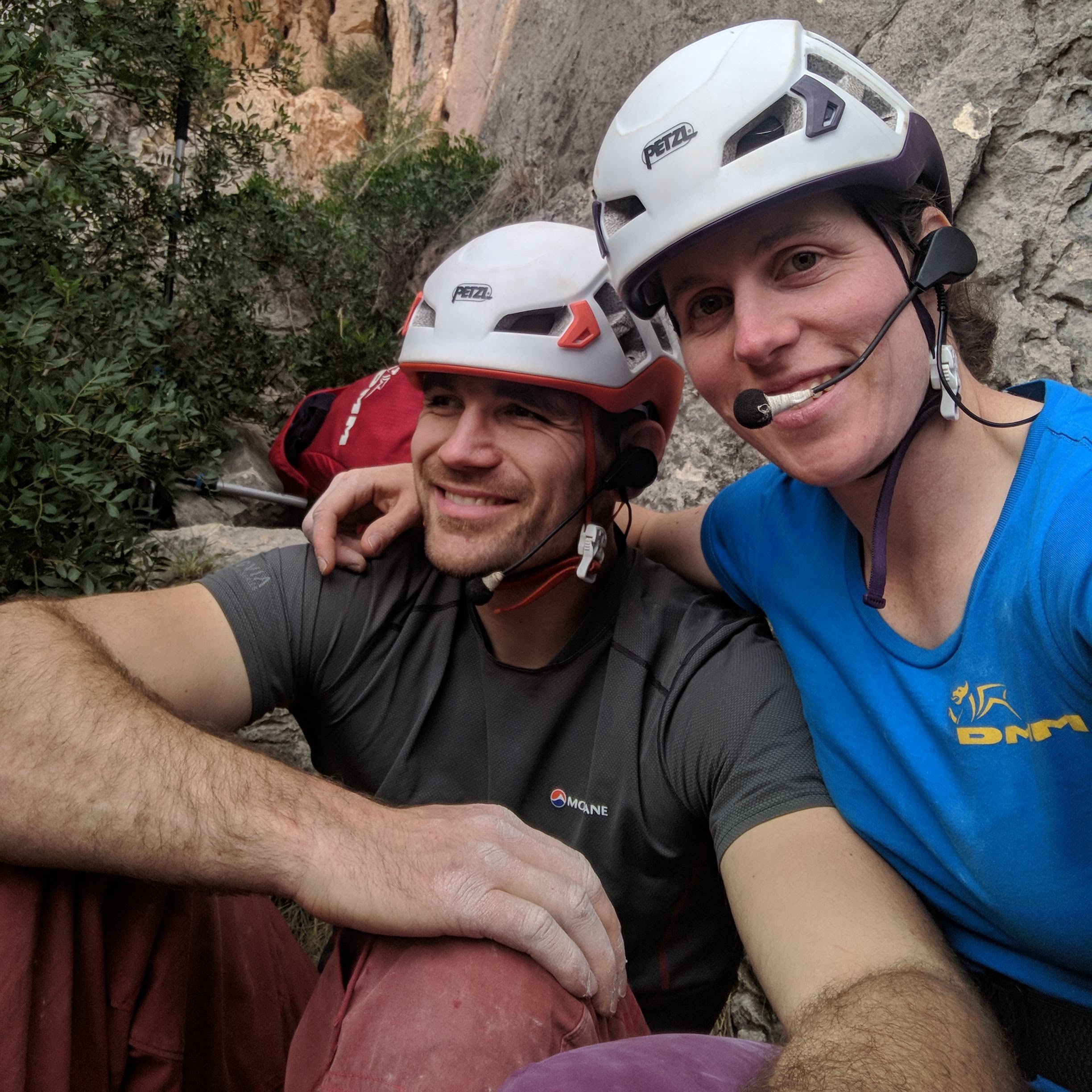A photo of Jesse and Molly with the helmets and headsets on, sitting at the base of a crag. They both have big smiles on their faces.