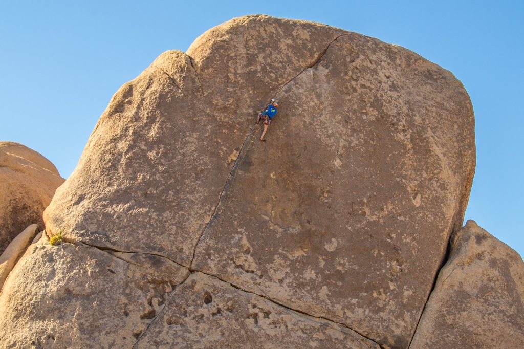 A photo of jesse climbing Rubicon, a thin crack line up another huge boulder. The route is in the shade with clear blue sky above.