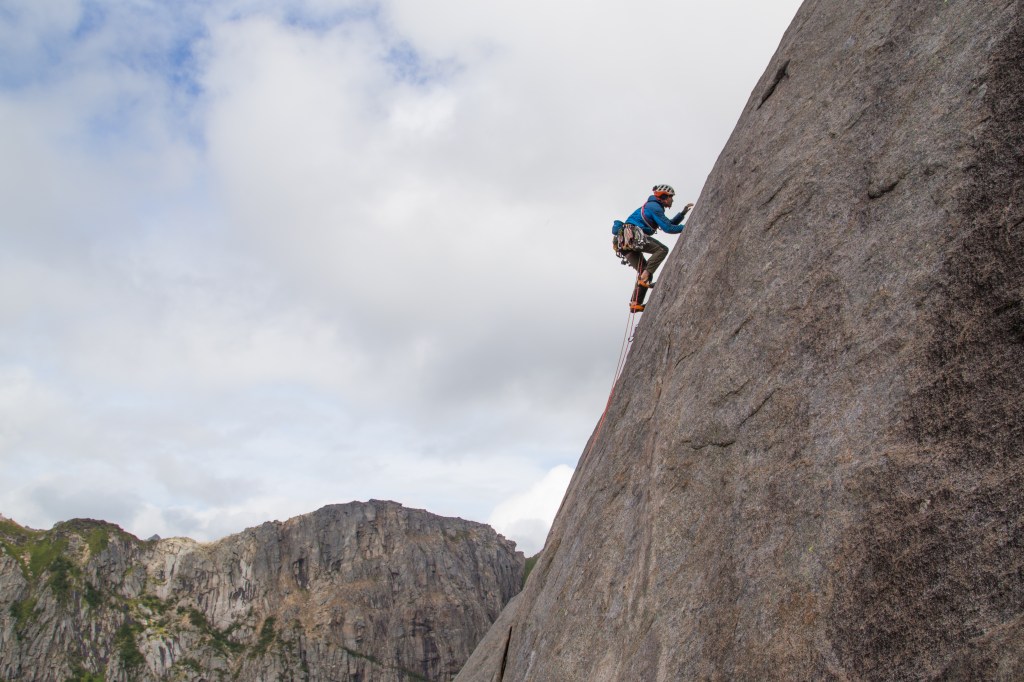 A photo of Jesse high on pitch 3 on Bare Blabaer, with some big spikey mountains in the background.