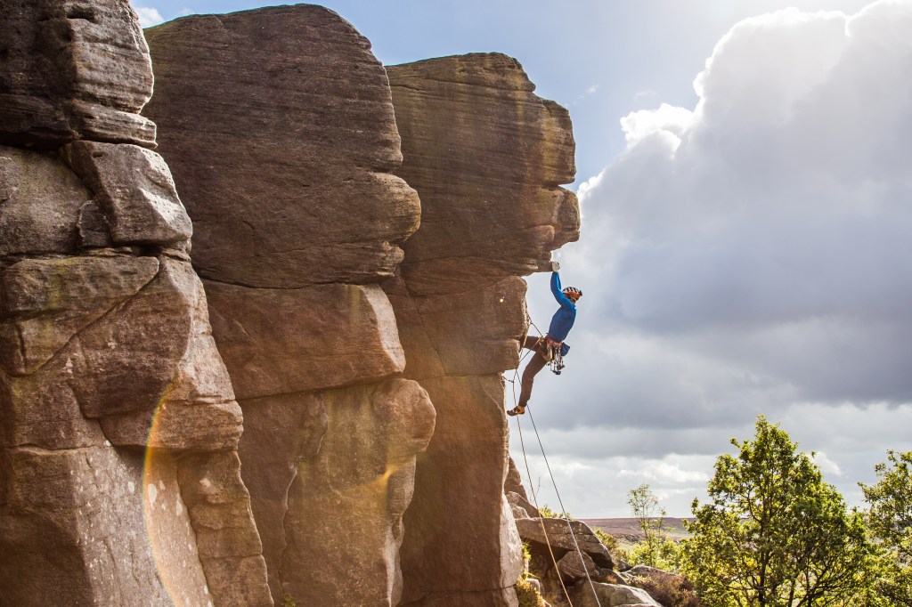 Jesse climbing the Sentinal E2 5b, Peak District, UK. The picture shows 3 large prows of rock jutting out stacked against each other. Jesse is hanging on with both hands on the hold from the further prow. He has gear placed by his waist and rock above his head. The next moves look steep and overhung. There are grey clouds in the sky and greens trees below him glistening in the sun.