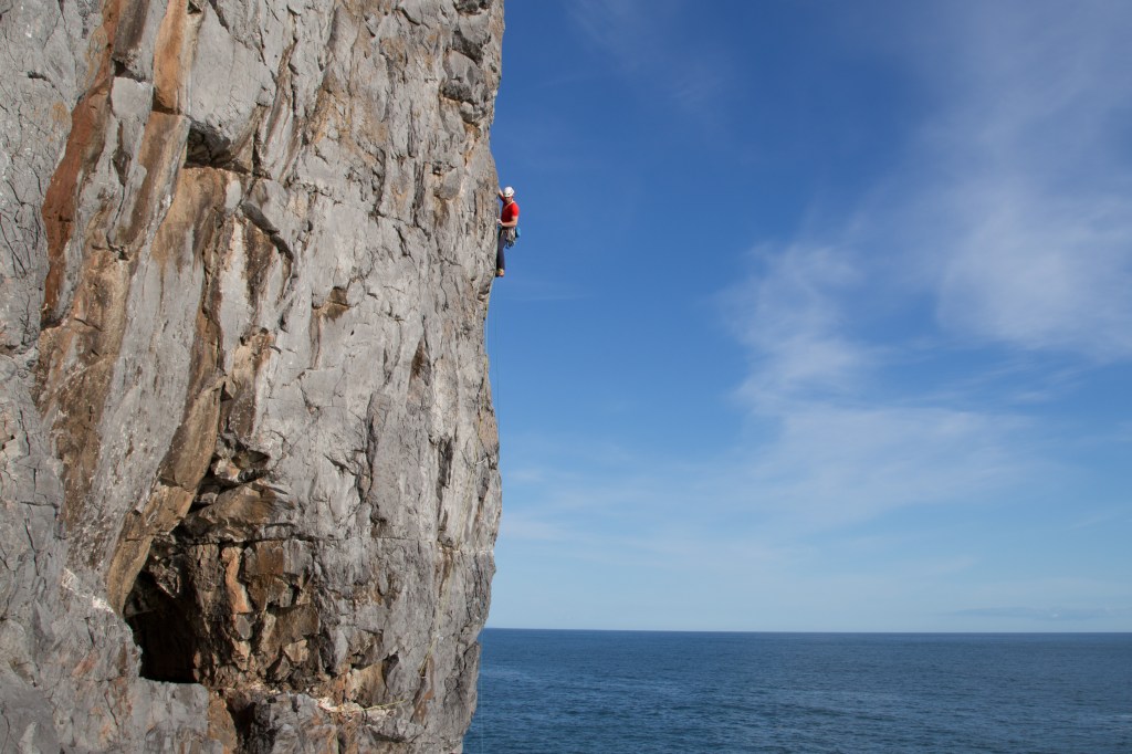 Jesse climbing Cool For Cats E1 5b, Pembroke, Wales. The picture shows a steep rock wall on the left hand side of the image and a beautiful blue sky and calm blue sea on the right. Jesse is high on the rock face silhouetted against the sky, he is reaching down with his left hand in search of a foothold. His feet are on a small edge and his right hand is high just out of view round the arete. His ropes dangle down below him and he stands out in a bright red t-shirt and white helmet.