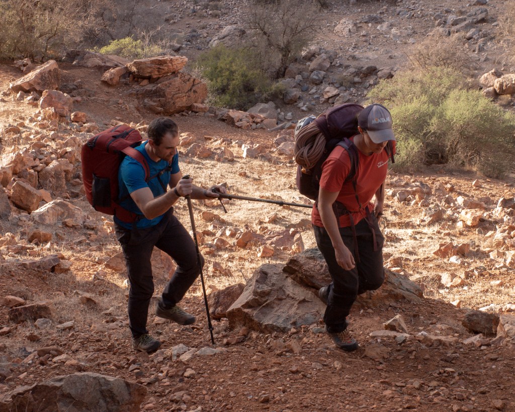 A photo showing Molly and Jesse walking on rocky uneven ground. Jesse is holding on to a walking pole that Molly is carrying, so he is able to follow behind.