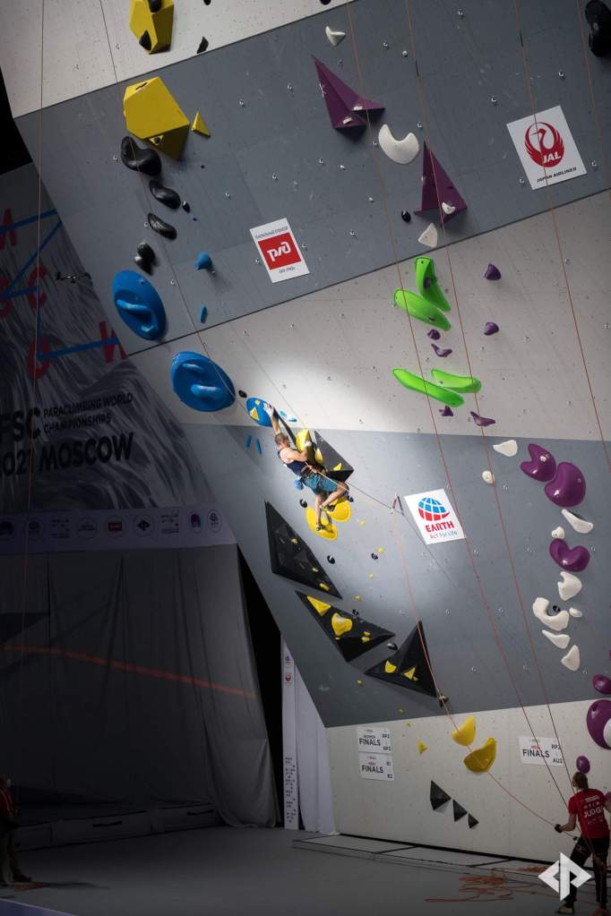 A photo showing Jesse in the spot light on a steep competition wall. He is reaching high with his left hand.