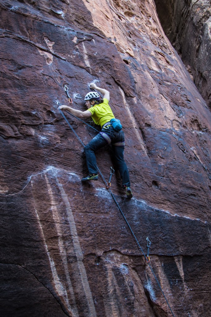 A photo of Molly clinging on for dear life with her right finger tips on a tiny crimp. She is hold the rope in her left hand, about a foot away from the quickdraw. She is transfixed, looking extremely concerned about not being able to make the clip. Her feet on are a narrow ledge.