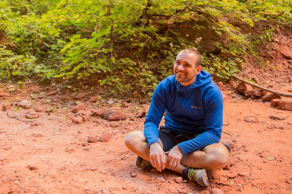A photo of Jesse sat on the ground, orange sand and small rocks. He is looking up with a big smile on his face, wearing a blue top and shorts. He looks like he needs a shower.