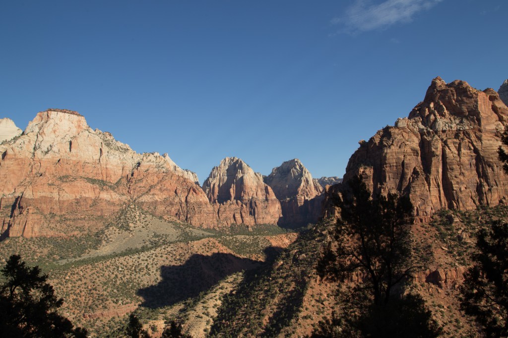 A photo showing the landscape in Zion National Park, clear blue sky with large rocky mountains in the background. The rock has different strata layers, the top being white, the middle orange and the lower part is dark red. All the mountains have horizontal stripes showing these different bands of colour.  