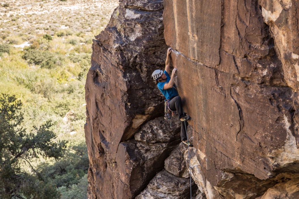 Molly climbing Left Out 5.10d Red Rock, USA. The picture shows molly in the centre of the image, climbing a steep wall. Her left hand is reaching high into a horizontal break and her right hand is crimping on a small edge at her eye level. Her right foot is stepped high into a small depression and her left foot is on a large ledge. She is wearing a blue t-shirt and black trousers. The sun is blazing in the background over the scrub-land, but the crag is in shade.