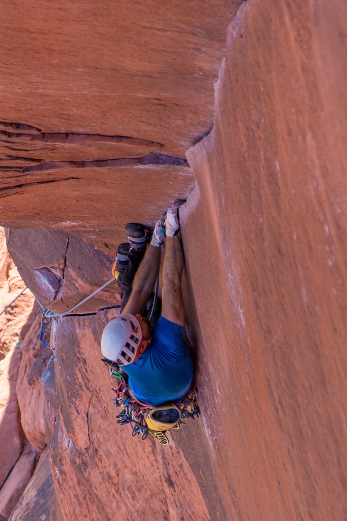 The photo shows Jesse in the layback position, with 2 hands pulling on the finger sized corner crack and 2 feet pushing against the left smooth wall. The position looks strenuous, he is carrying many cams on his harness and is wearing a bright blue t-shirt which shows up well against the red rock.