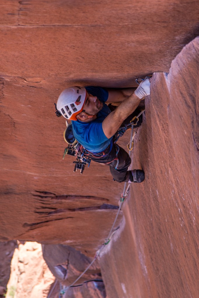 A photo showing Jesse placing a cam with his right hand as high as he can reach in the corner crack. His left hand and left foot are jammed in the crack and his right foot is smeared against the vertical rock face on the right side of the corner.