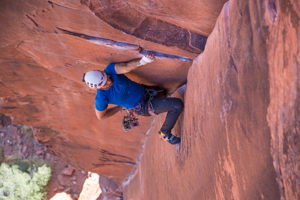 Jesse climbing The Fox 5.10d, Red Rock USA. The picture shows Jesse in the center of the image, hanging onto a small ledge with his left hand on the left side of large soaring corner. He is dipping for chalk with his right hand. Large cams hang from his harness and his right foot is perched on a tiny edge on the right wall and he is leaning back with a straight arms. It looks like a strenuous position but his face looks calm and chilled.