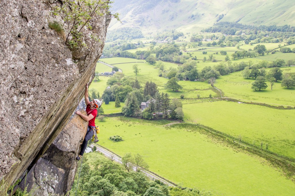 Jesse climbing The Bludgeon E1, Borrowdale, Lake District. The picture shows an overhanging rock wall on the left and a luscious green area of fields and trees in the valley below on the right. Jesse is wearing a red t-shirt and is hugging a rock spike with his left arm, while reaching high with his right hand. The climb looks quite improbable from this angle as it is so steep. The radio ear piece is visible in his left ear.