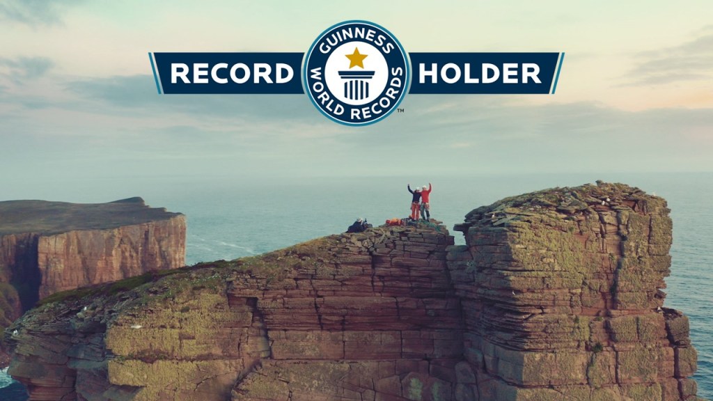 A picture of Molly and Jesse on top of the Old Man of Hoy with arms out stretched. There is a banner across the top of the picture with the guinness world records logo and record holder written next to it.