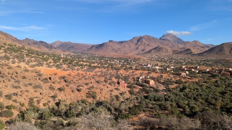 A view across the desert like landscape to the mountains in the background. There are houses and a minaret in the mid distance and the sun is baking the ground.